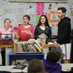 four middle school students stand at the front of a classroom to talk to fifth graders