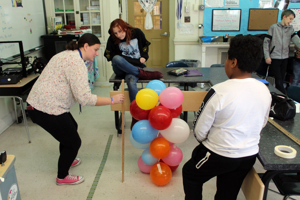 teacher measuring baloon tower while girl and boy look on