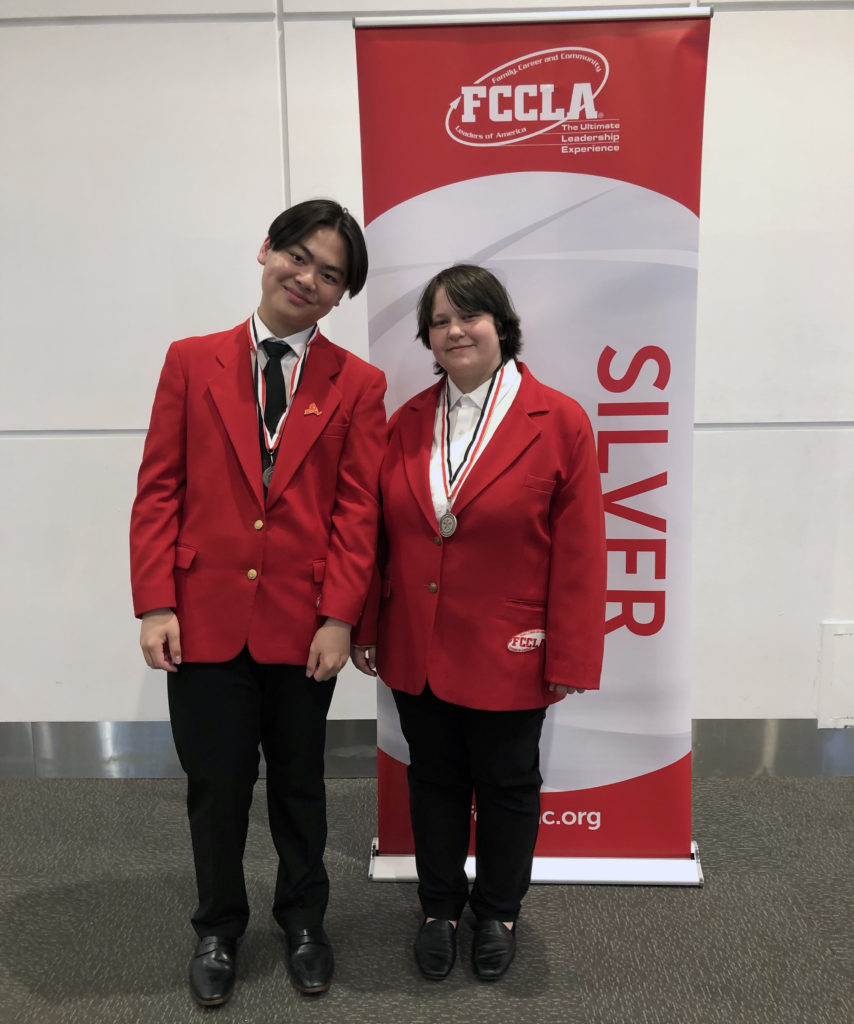 two students pose in red blazers next to silver sign