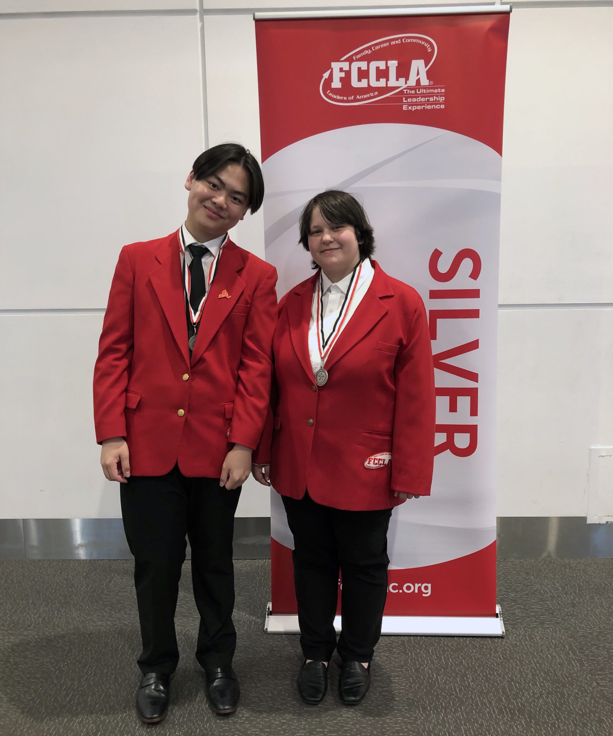 two students pose in red blazers next to silver sign