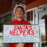 Student wearing Santa hat and holding Santa's Helpers sign.