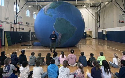 man standing in front of giant Earth balloon and student audience in gym.