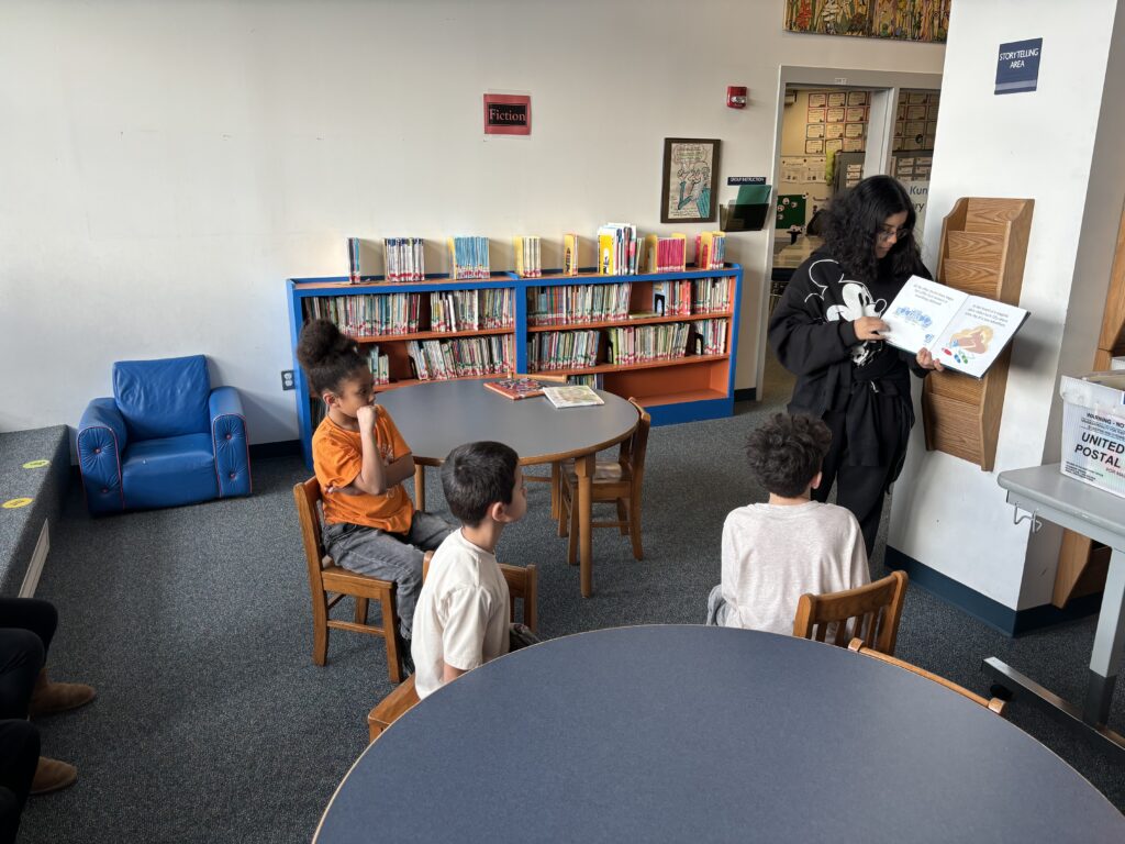 high school student reading group of elementary school students sitting at library tables.