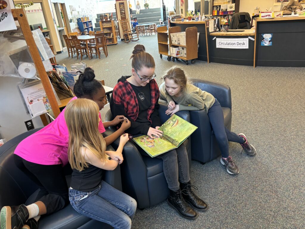 high school student reading group of elementary school students sitting on chairs.