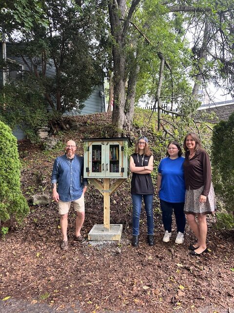 four people posing with tiny library consisting of wooden box with glass doors on post